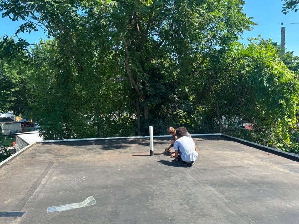 Person crouching on a rooftop near a white vent, with green trees and blue sky in the background