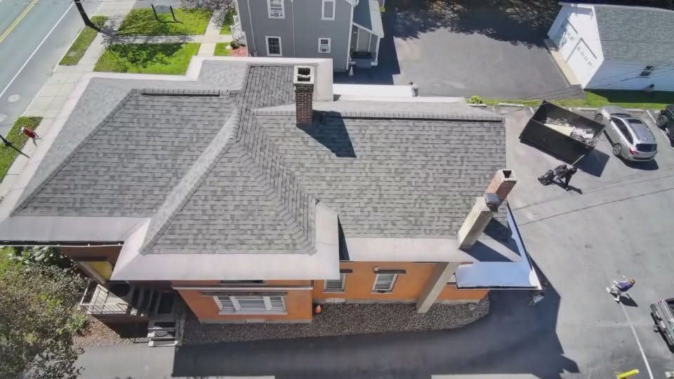 Aerial view of a small brick building with a gray shingled roof beside a parking lot and street.