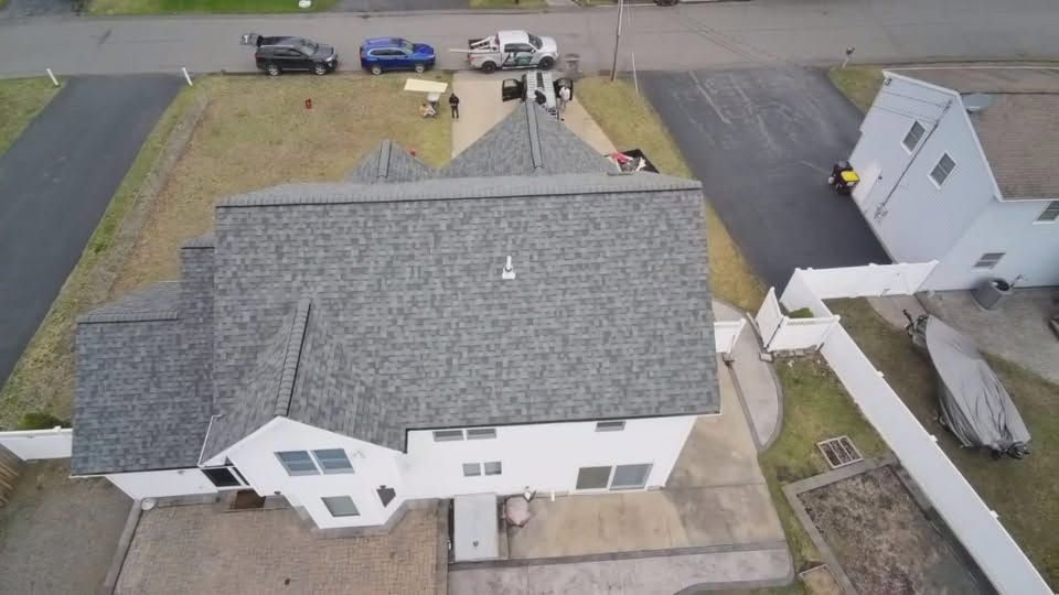 Aerial view of a house with gray roof, driveway, and nearby parked vehicles in a residential neighborhood