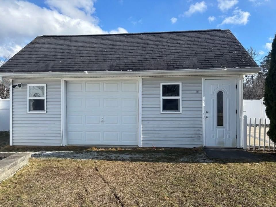 Small white house with gray roof, garage door, front door, and a grassy yard under a blue sky