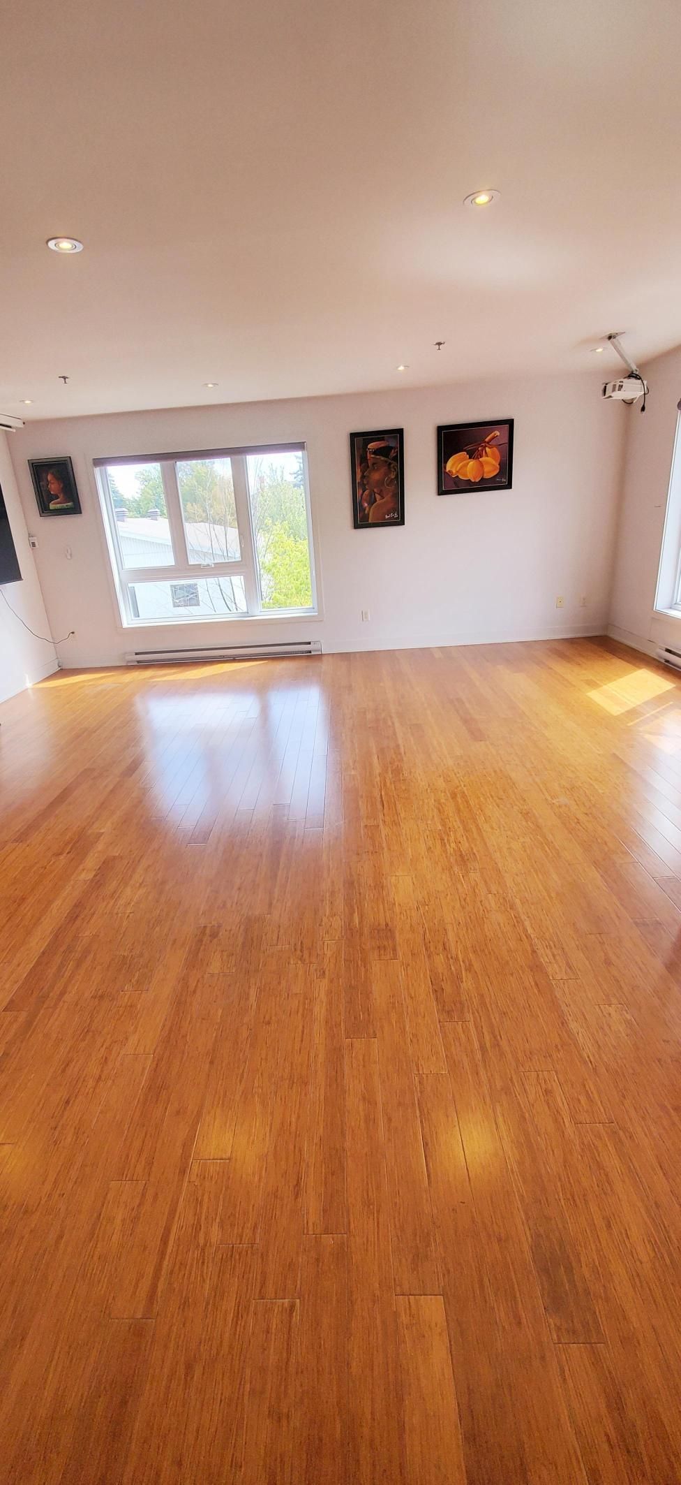 Chambre vide avec parquet, murs blancs, fenêtres et œuvres d'art encadrées.