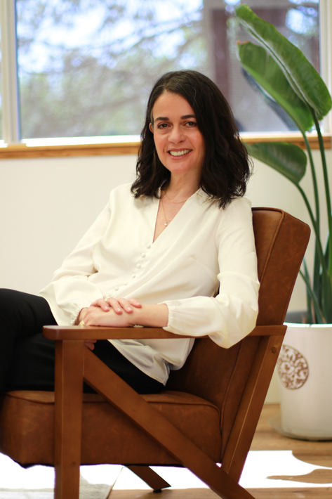Woman with dark hair smiles, sitting in a brown armchair, wearing a white blouse. She is indoors with a window and a potted plant.