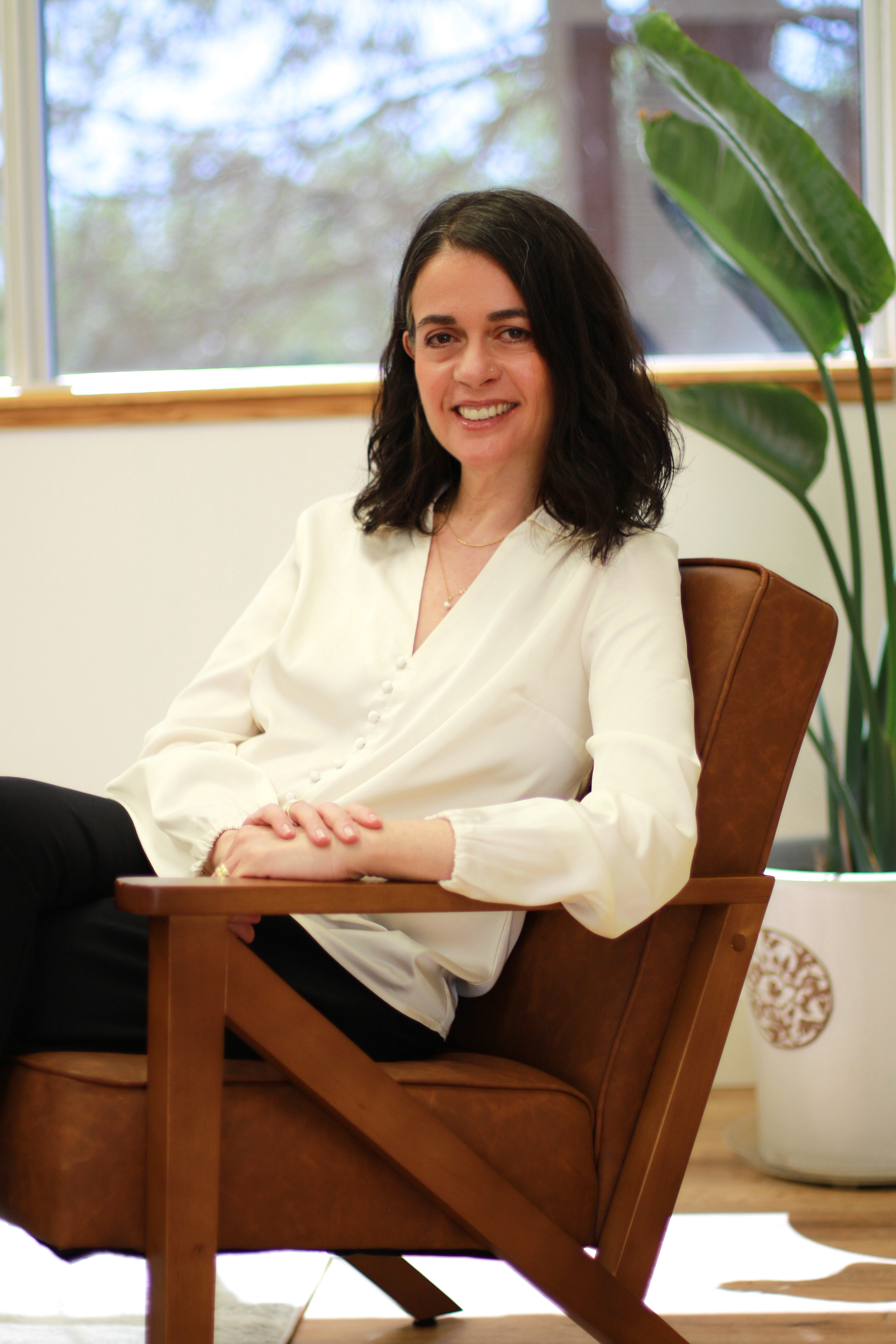 Woman with dark hair smiles, sitting in a brown leather armchair indoors. She wears a white blouse and black pants, in a well-lit room.