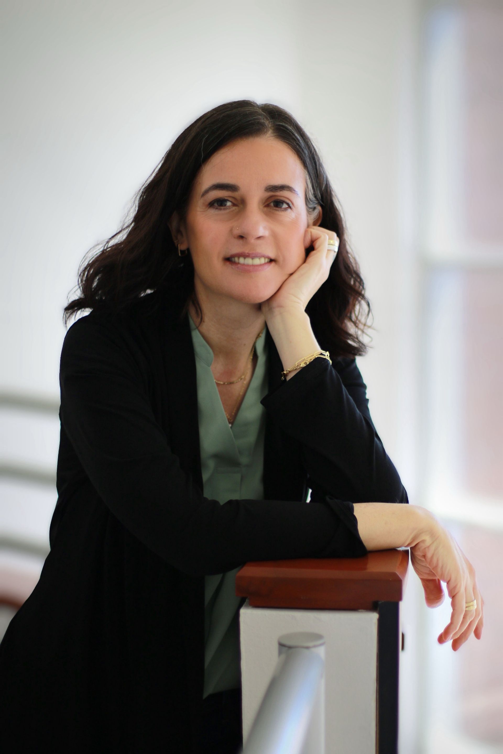 Woman in a black blazer and light green shirt leans on a railing, smiling at the camera.