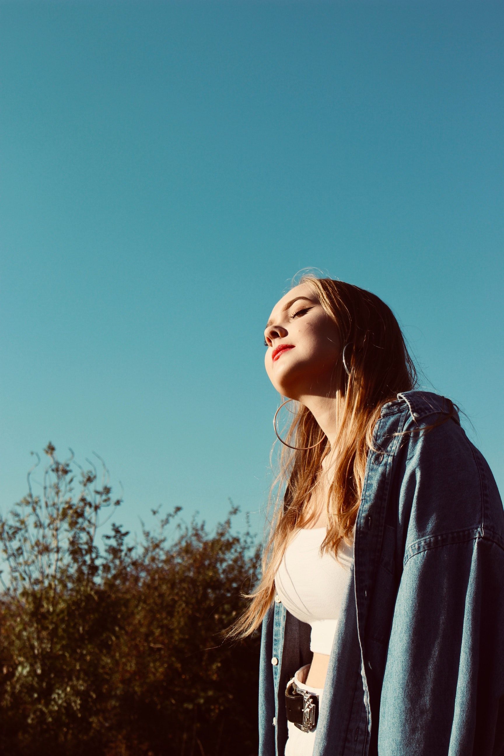 Woman in denim jacket looks up at blue sky, bathed in sunlight. Blonde hair, hoop earrings.