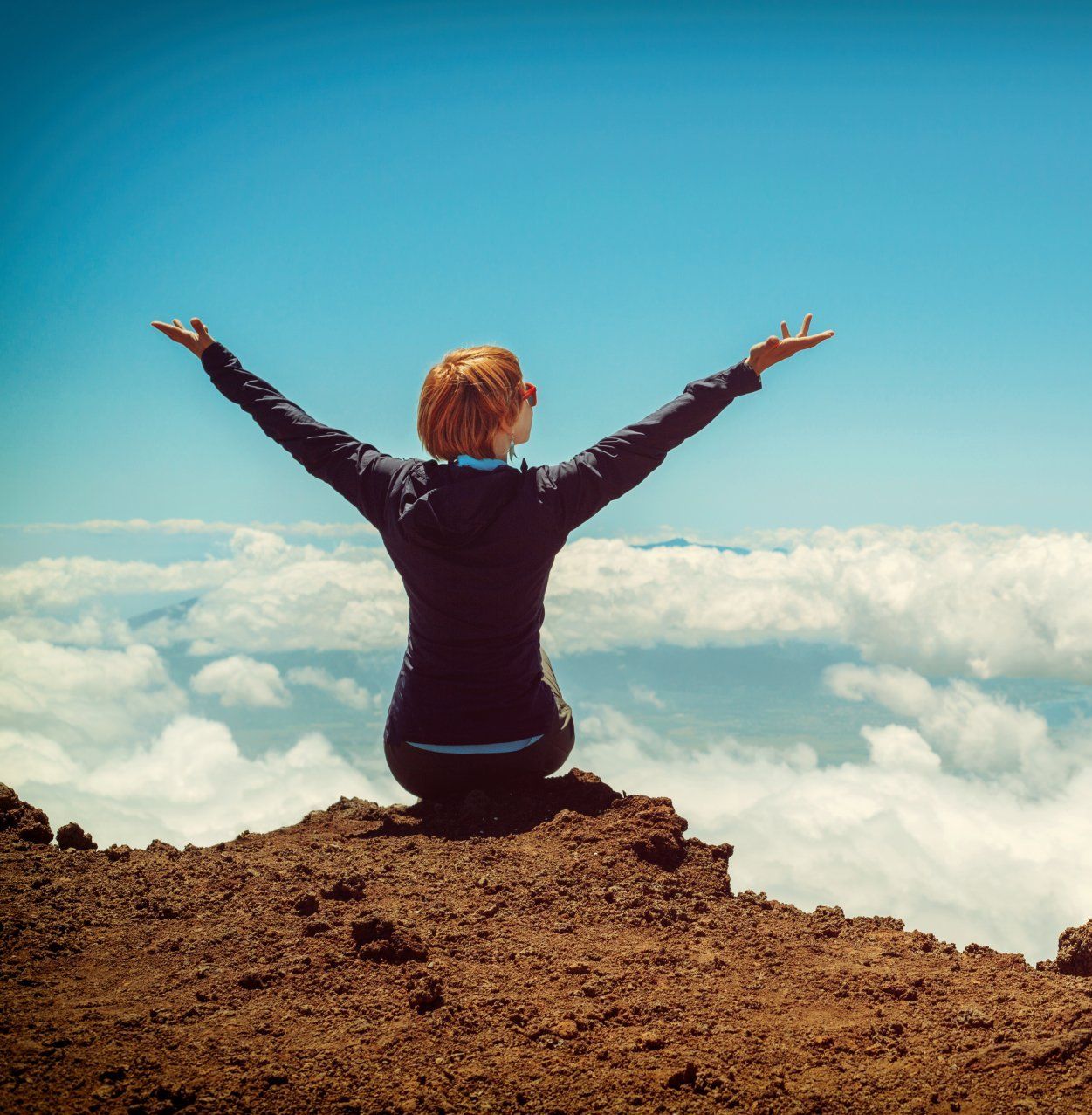 Woman with arms outstretched, seated on a mountaintop above clouds, enjoying a sunny day.