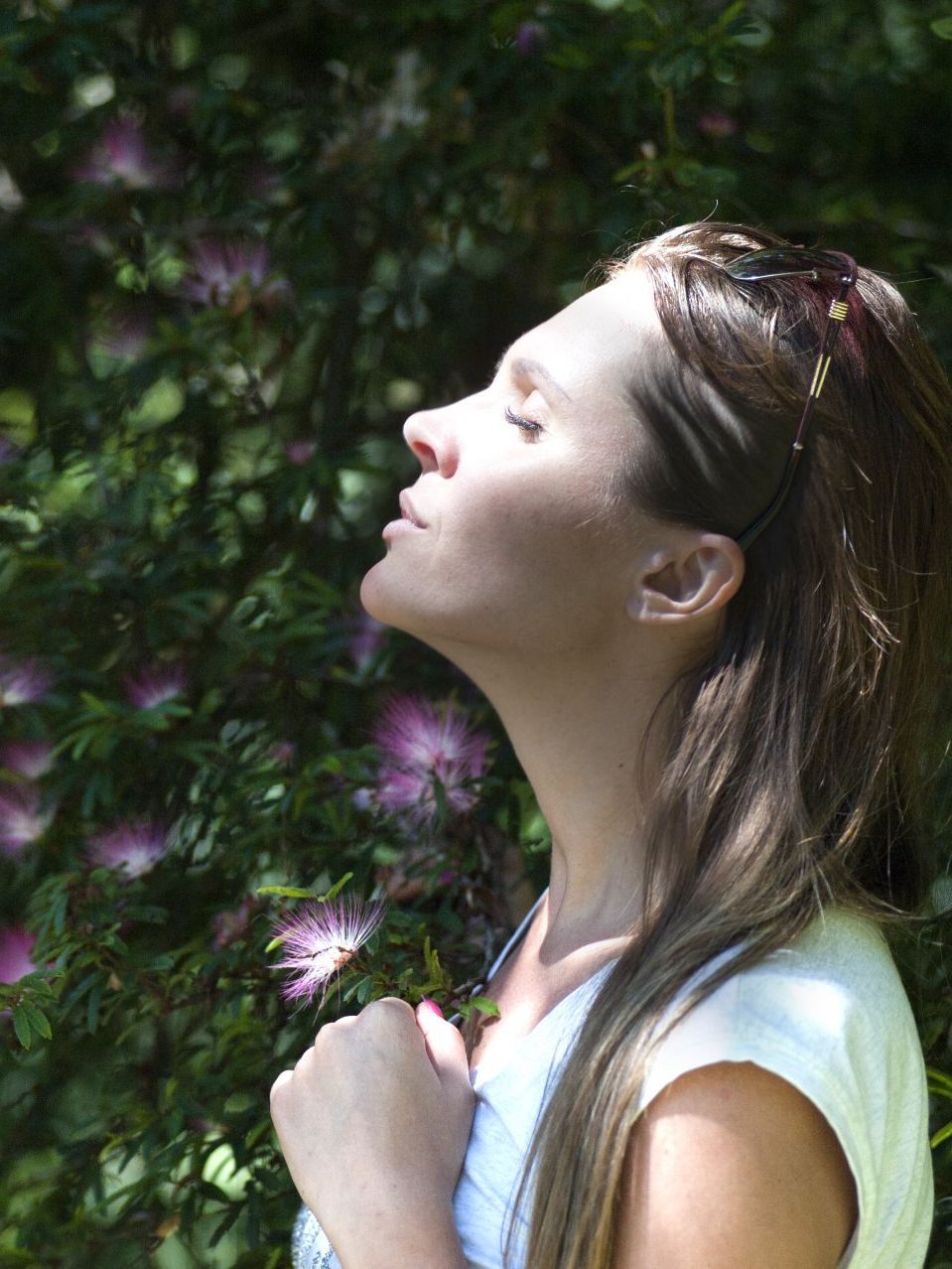 Woman with eyes closed, face tilted up, enjoying sunlight in a garden setting. Long hair, white top, hands clasped.