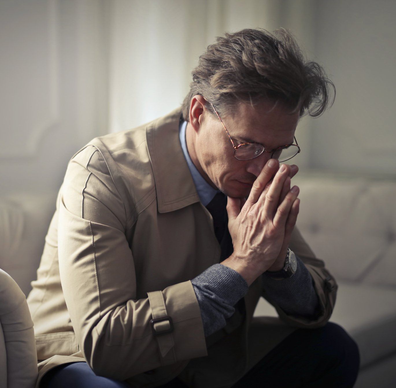 Man in glasses and tan coat, sitting with hands clasped in front of him, looking down with a concerned expression.