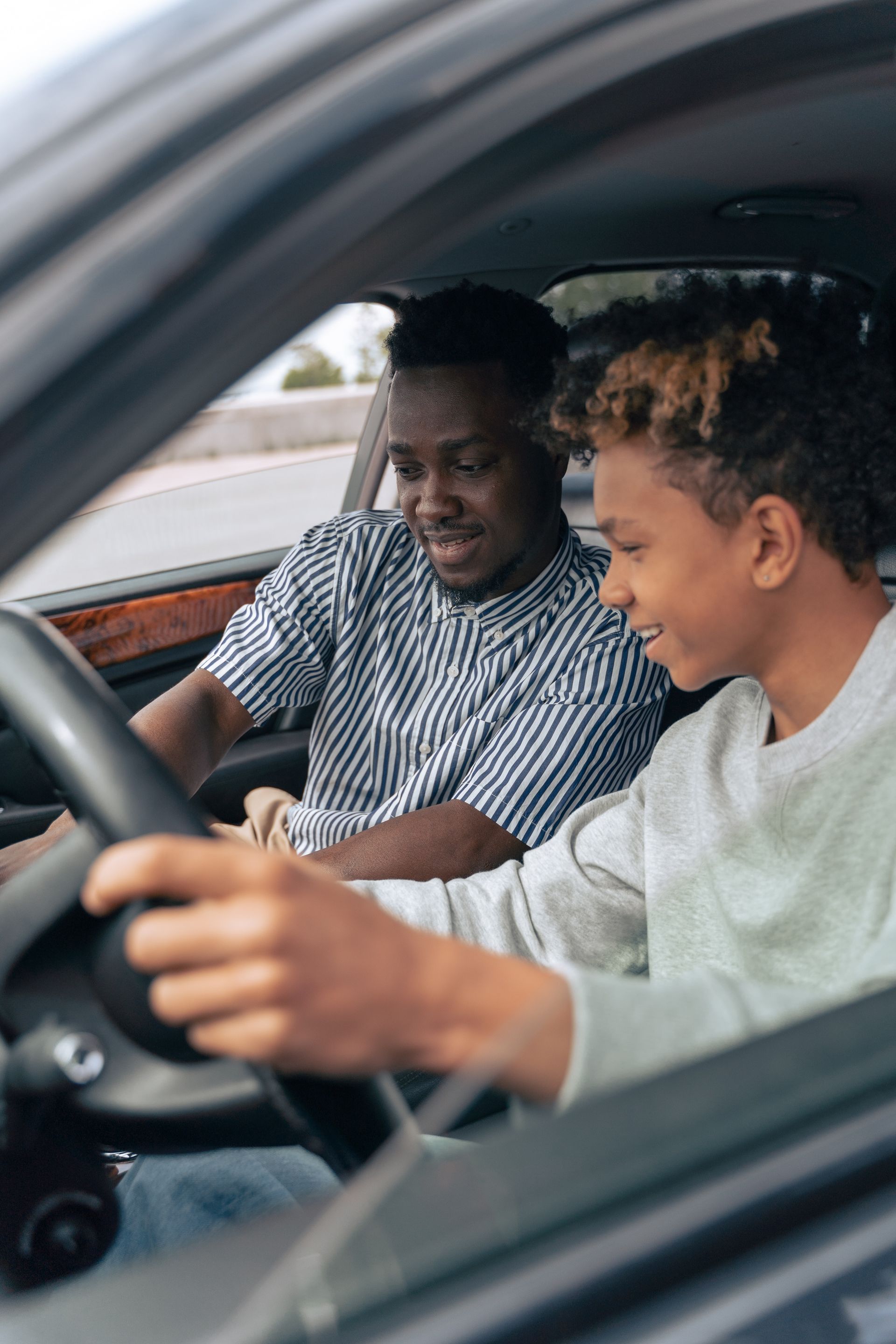 A young man learns to drive with guidance from an older man in a car. The young man holds the steering wheel and smiles.