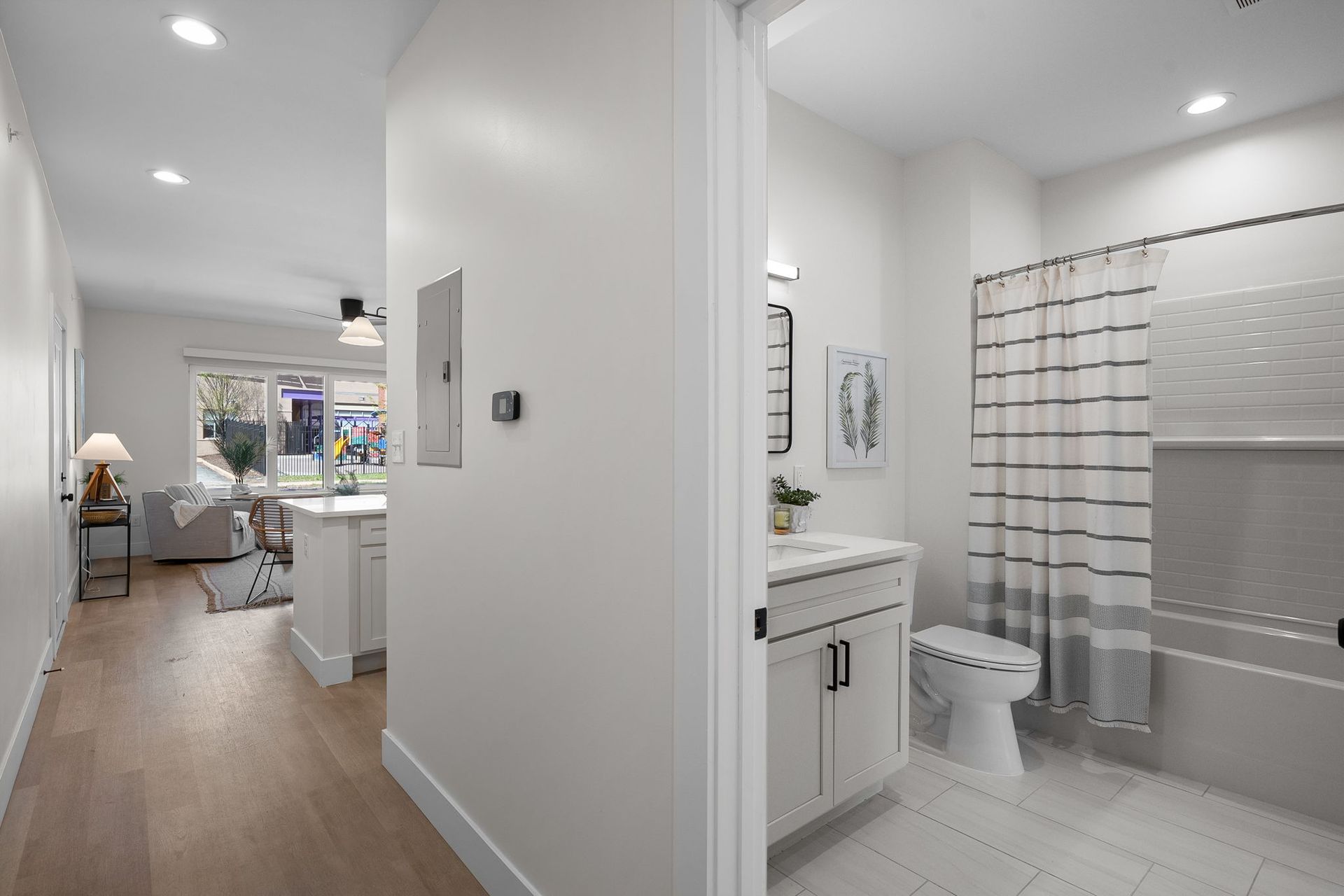 Bright hallway beside a bathroom with gray shower curtain, leading to a sunlit living room and kitchen