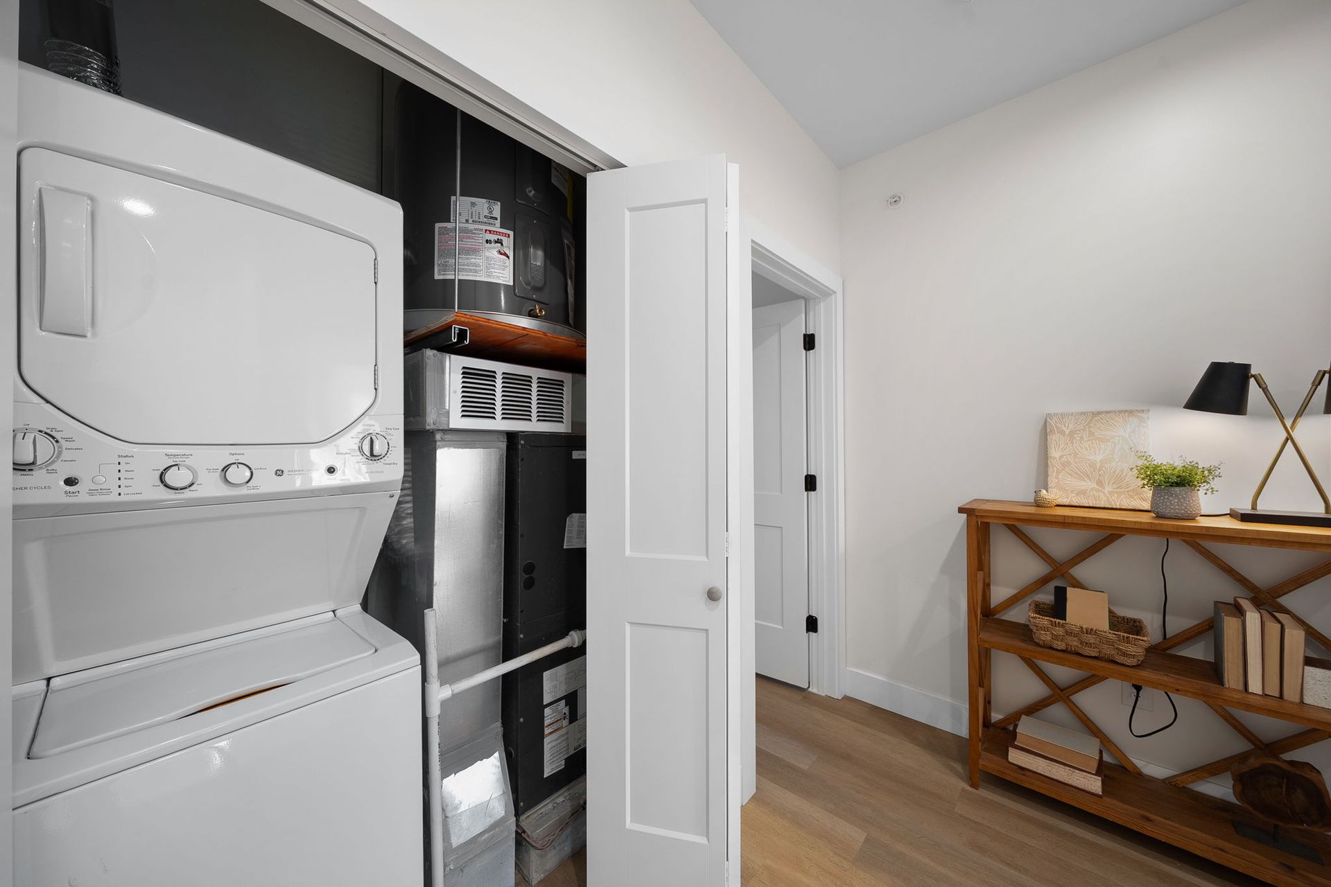 Laundry closet with stacked washer and dryer beside a hallway and wooden shelving unit