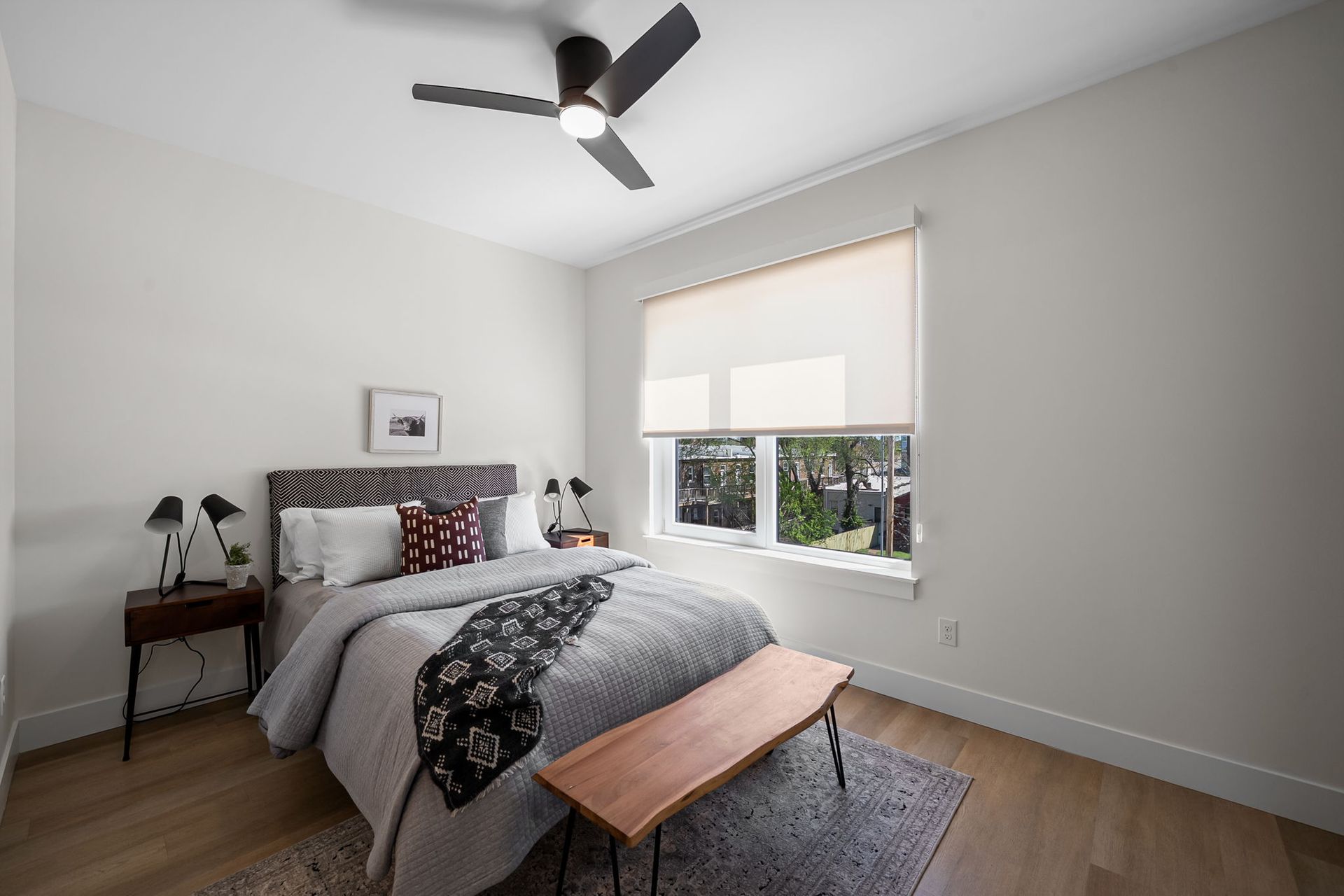 Modern bedroom with gray walls, bed, bedside table, ceiling fan, and window with blinds.