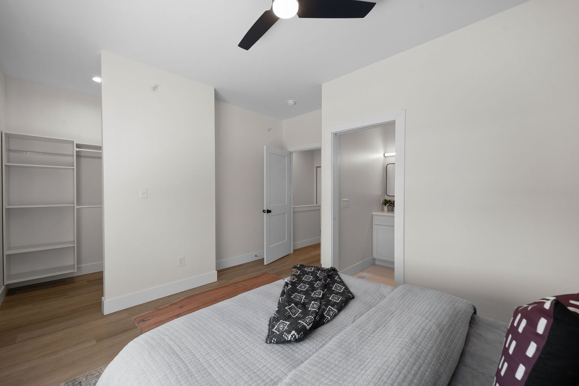 Bright bedroom with gray bed, open closet, white walls, hardwood floor, and ceiling fan