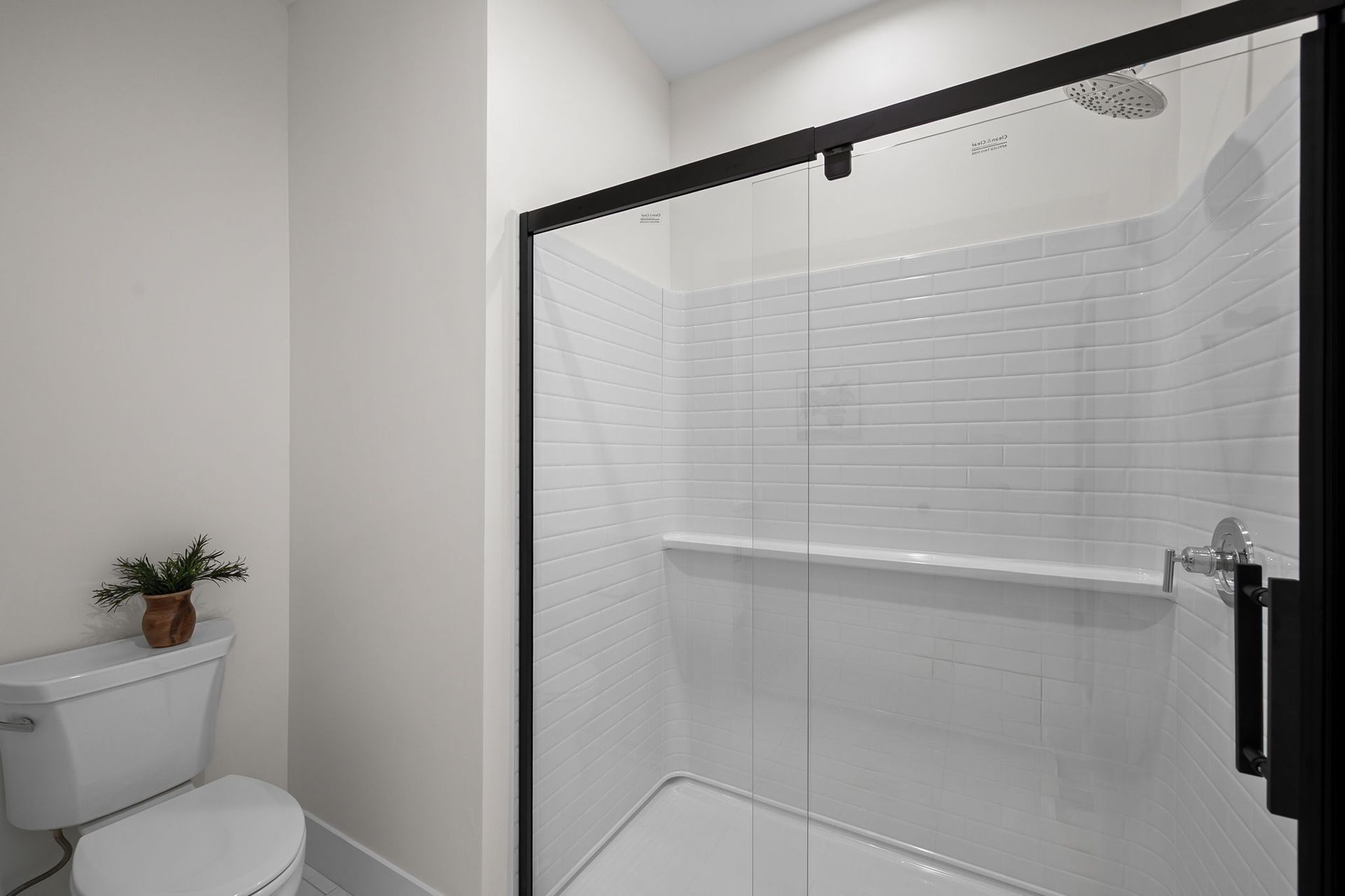 Modern bathroom with glass shower, white tile, toilet, and a small plant on the tank