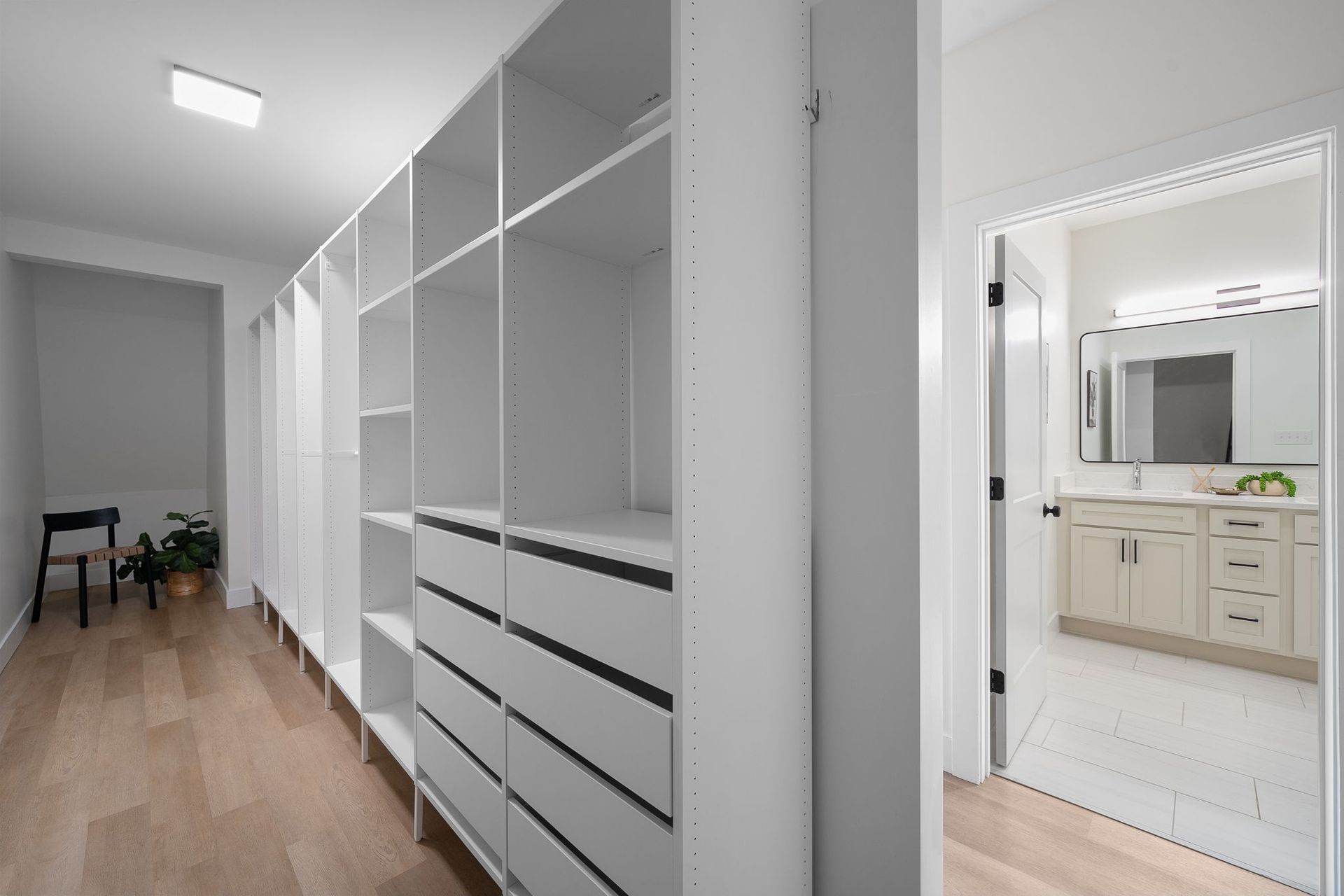 Bright white walk-in closet with built-in shelves and drawers, opening to a bathroom with a vanity.