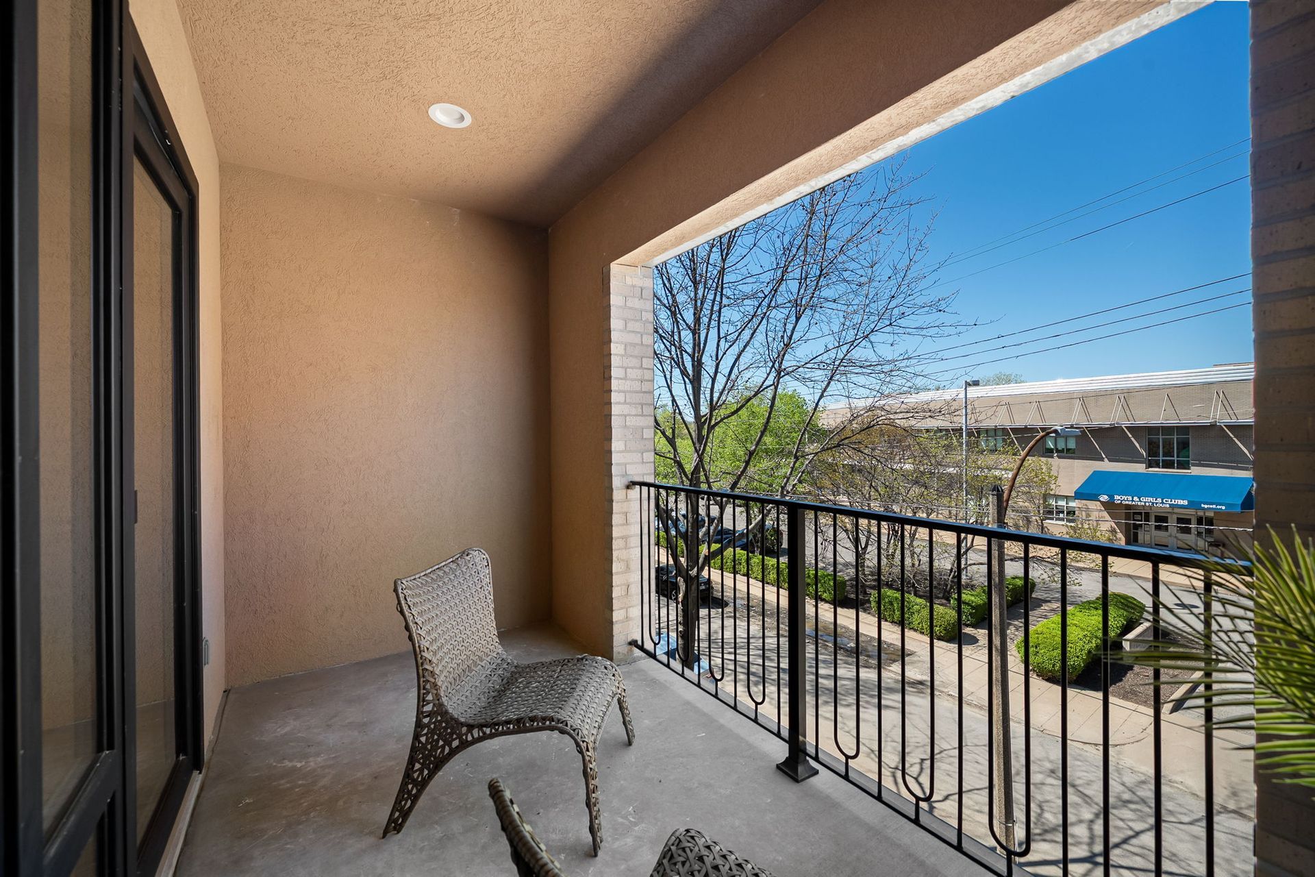 Apartment balcony with a chair, black railing, and view of trees and a pool on a sunny day