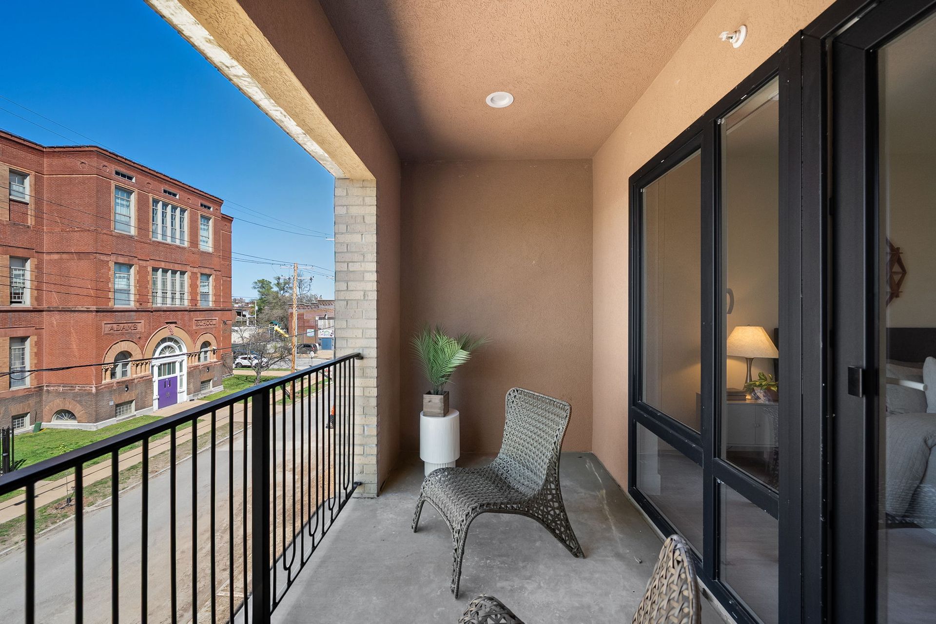 Apartment balcony with wicker chair, potted plant, black railing, and brick buildings across the street