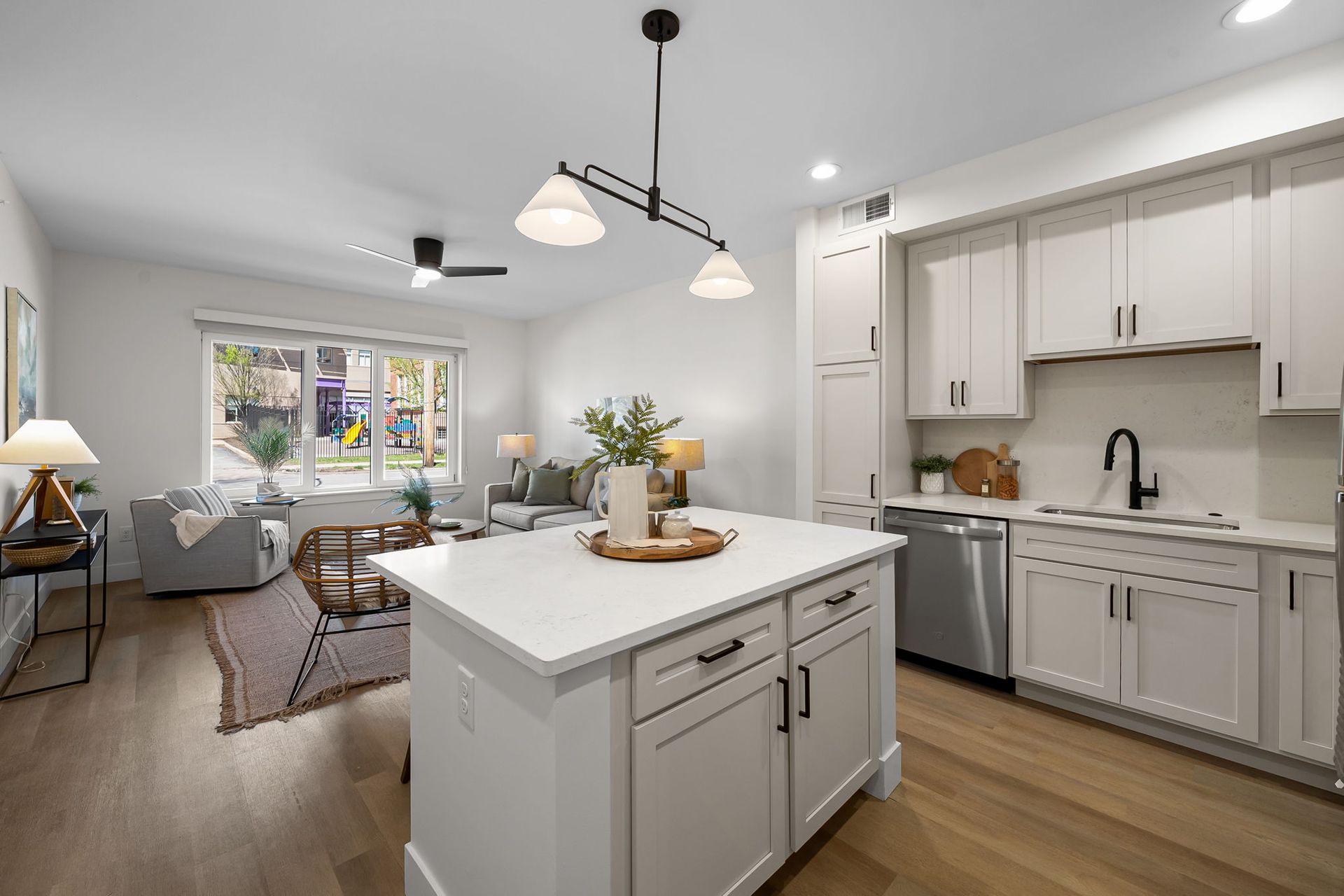 Bright open kitchen with white cabinets and island, leading to a cozy living area with sofa and large window.