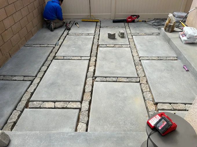 Worker laying concrete pavers in a patio beside a wall, with tools and materials nearby.