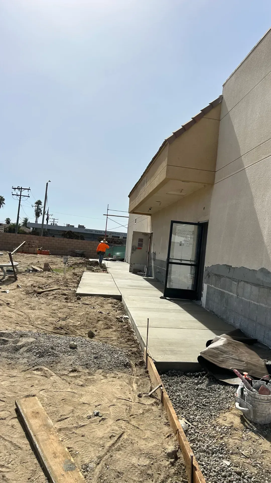 Building under construction; concrete walkway, worker in orange vest, dirt and sky visible.