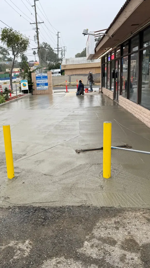 Workers smoothing wet concrete sidewalk outside a store on an overcast day. Yellow posts are visible.