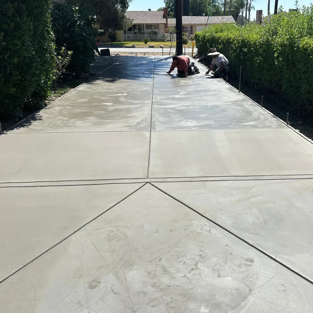 Workers smoothing fresh concrete on a driveway. Green bushes frame the scene; a house in the background.