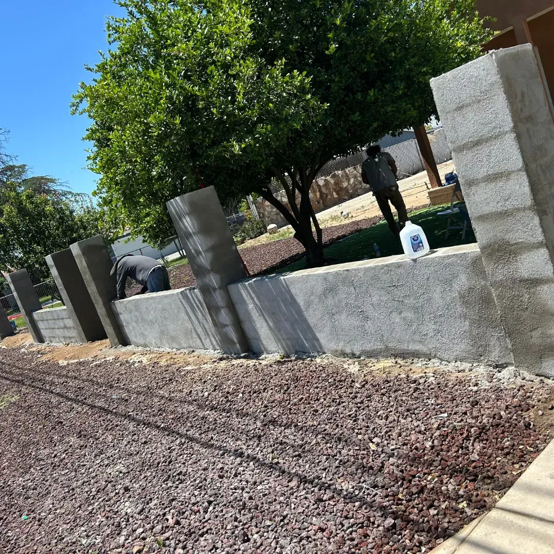 Concrete block wall construction with people working, trees, and blue sky.
