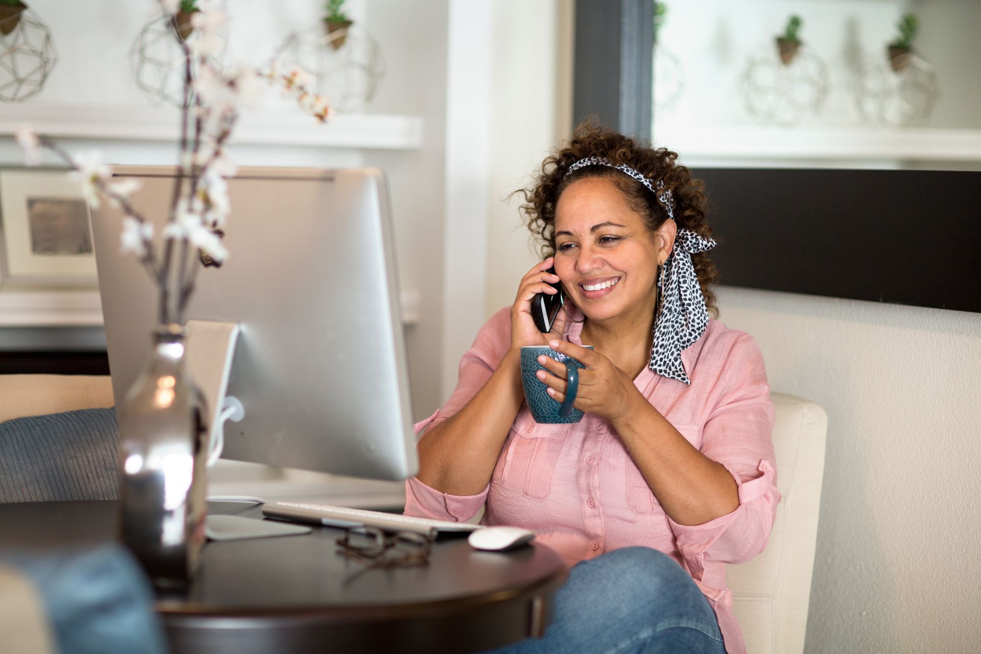 A woman is sitting at a table talking on a cell phone while holding a cup of coffee.