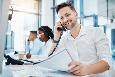 A man is sitting at a desk talking on a phone while holding a piece of paper.