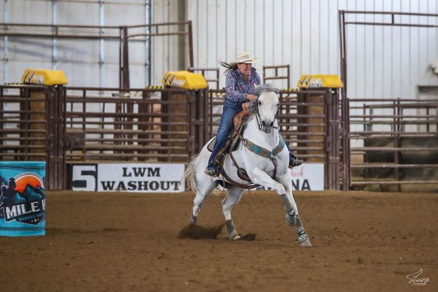 Cowgirl on a white horse turning in an indoor rodeo arena, with barrel and sponsor signs in the background
