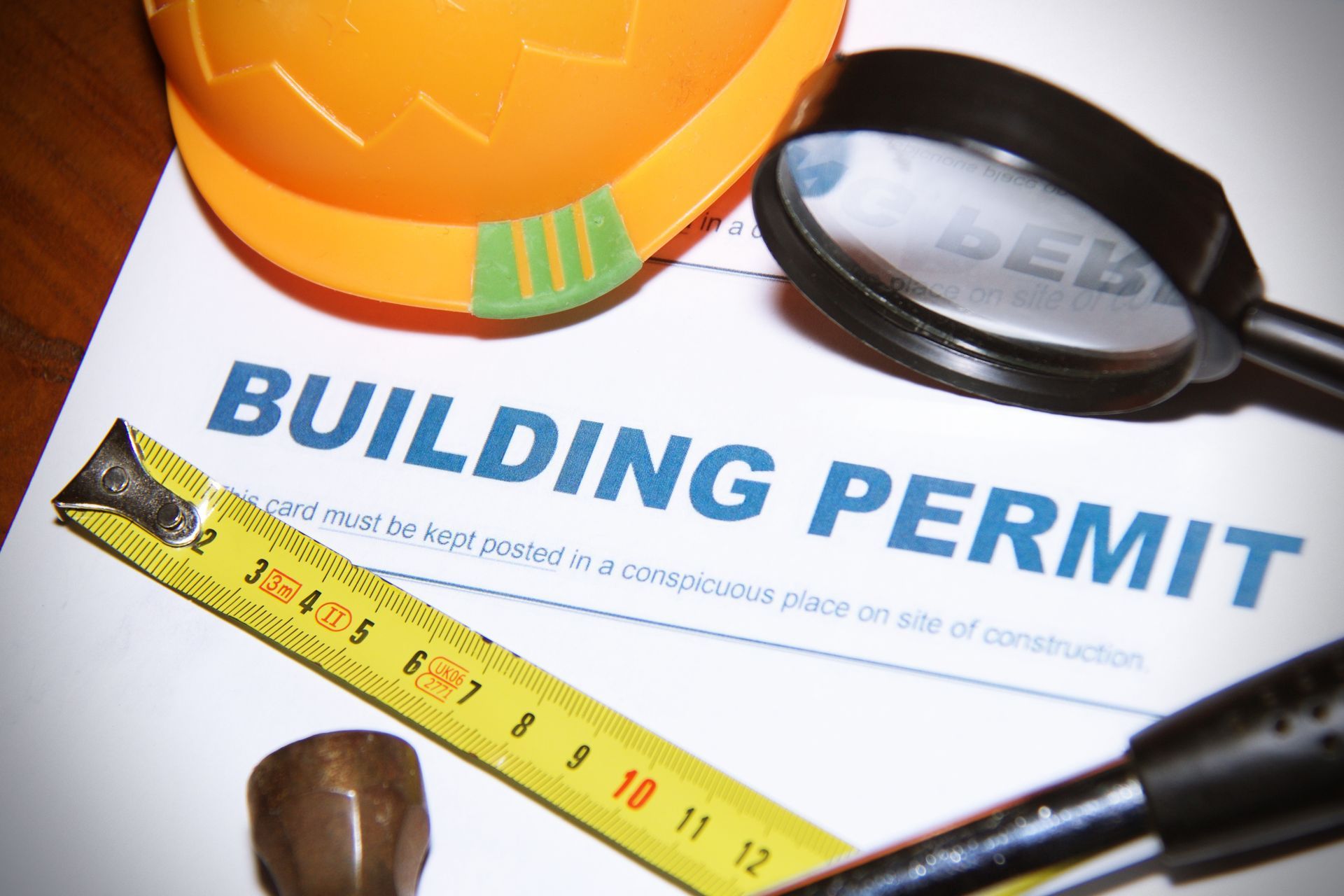 A building permit paper is surrounded by a yellow tape measure, a magnifying glass, and an orange hard hat.