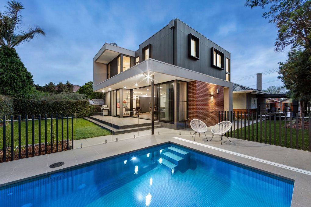 Modern two-story house with dark gray exterior, large glass walls, swimming pool, and patio at twilight.