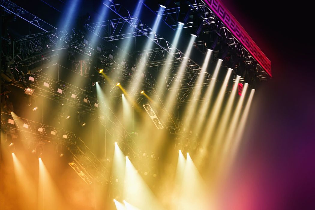 Le luci del palco proiettano fasci di luce bianca e dorata attraverso la fumosa scena del concerto.