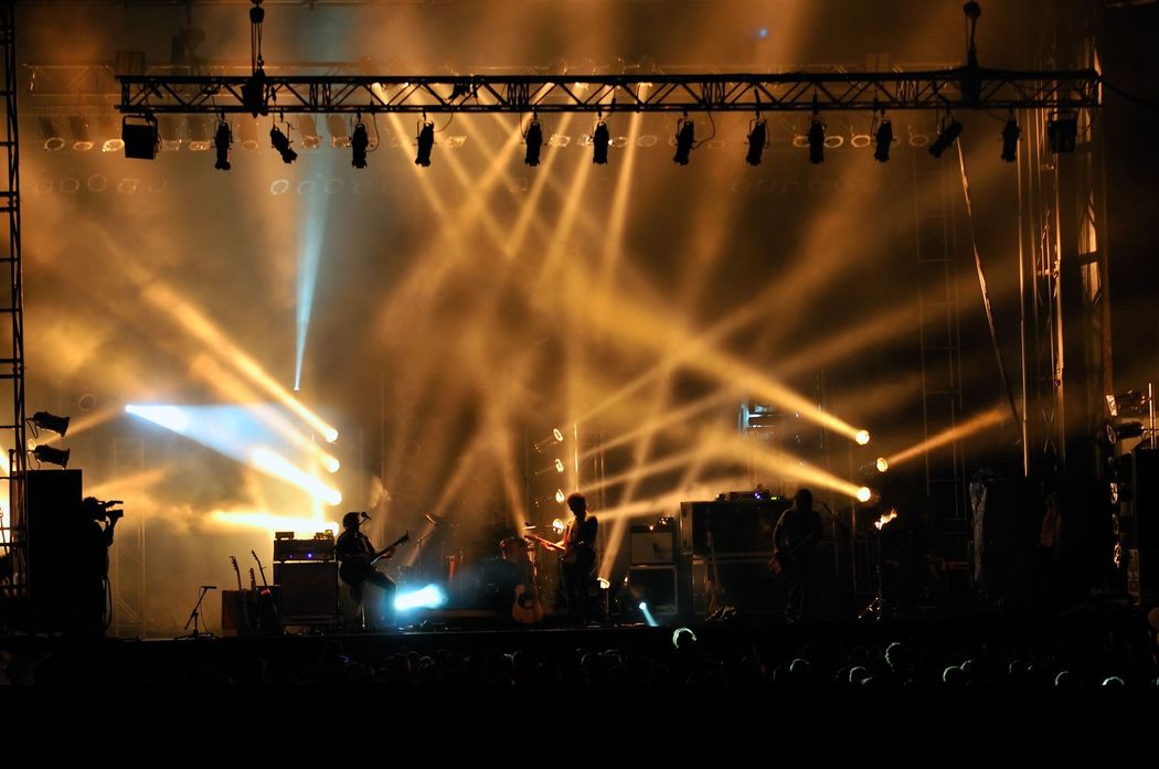 Palco da concerto illuminato da brillanti riflettori ambrati e musicisti in silhouette che si esibiscono di notte.