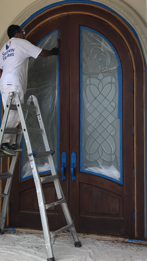 Person on ladder painting a dark wood double door, masking the glass panes with blue tape.