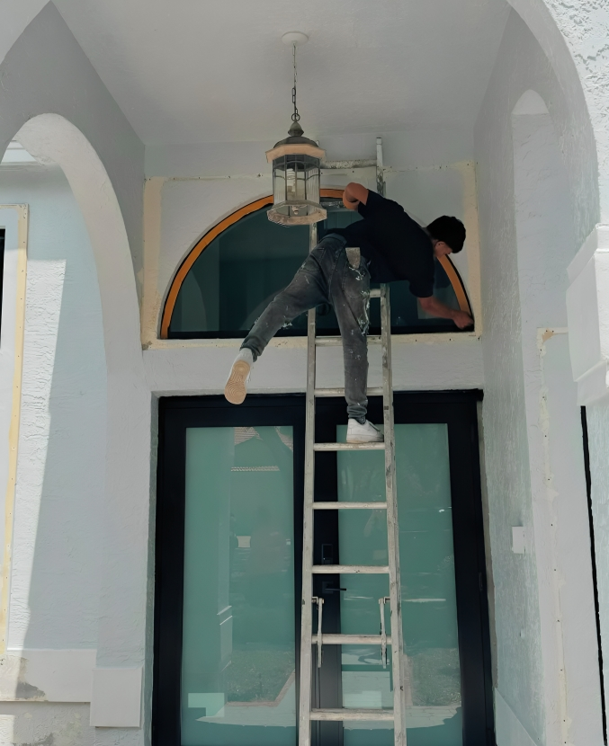 Man on ladder working near a large window, reaching up. Exterior setting, blue walls, archway.