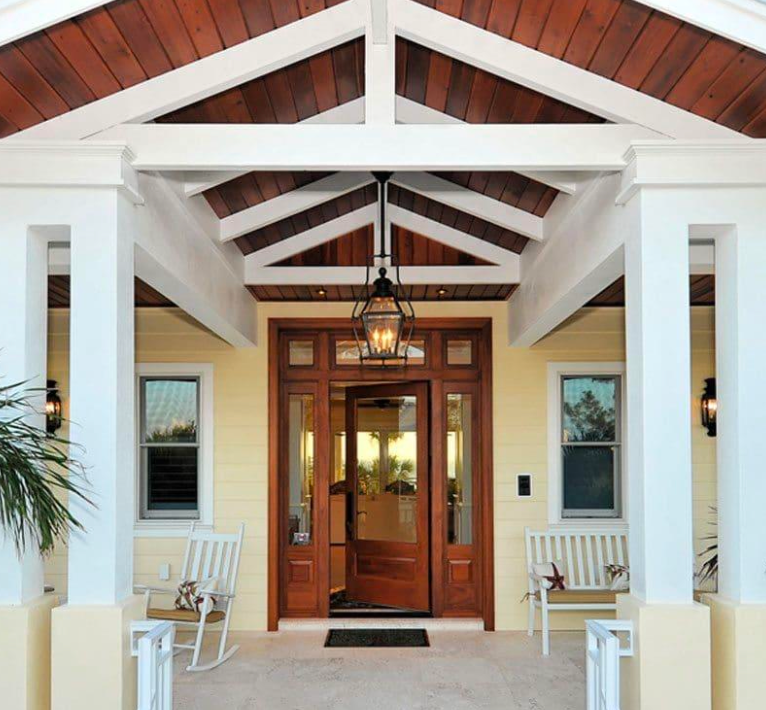 Yellow home entrance with brown wood door and white pillars. A bench and rocking chair flank the entrance.