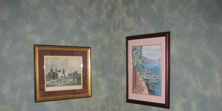 Two framed pictures on a mottled blue wall. One features a group of figures, the other a landscape.