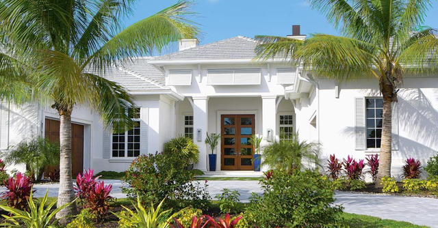 White house with wooden door, palm trees, and colorful plants under a blue sky.