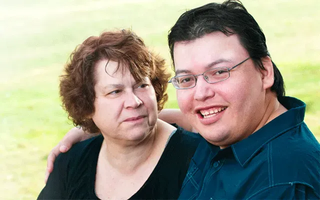 Two people sitting closely outdoors; one looks forward with a smile, while the other looks toward them.