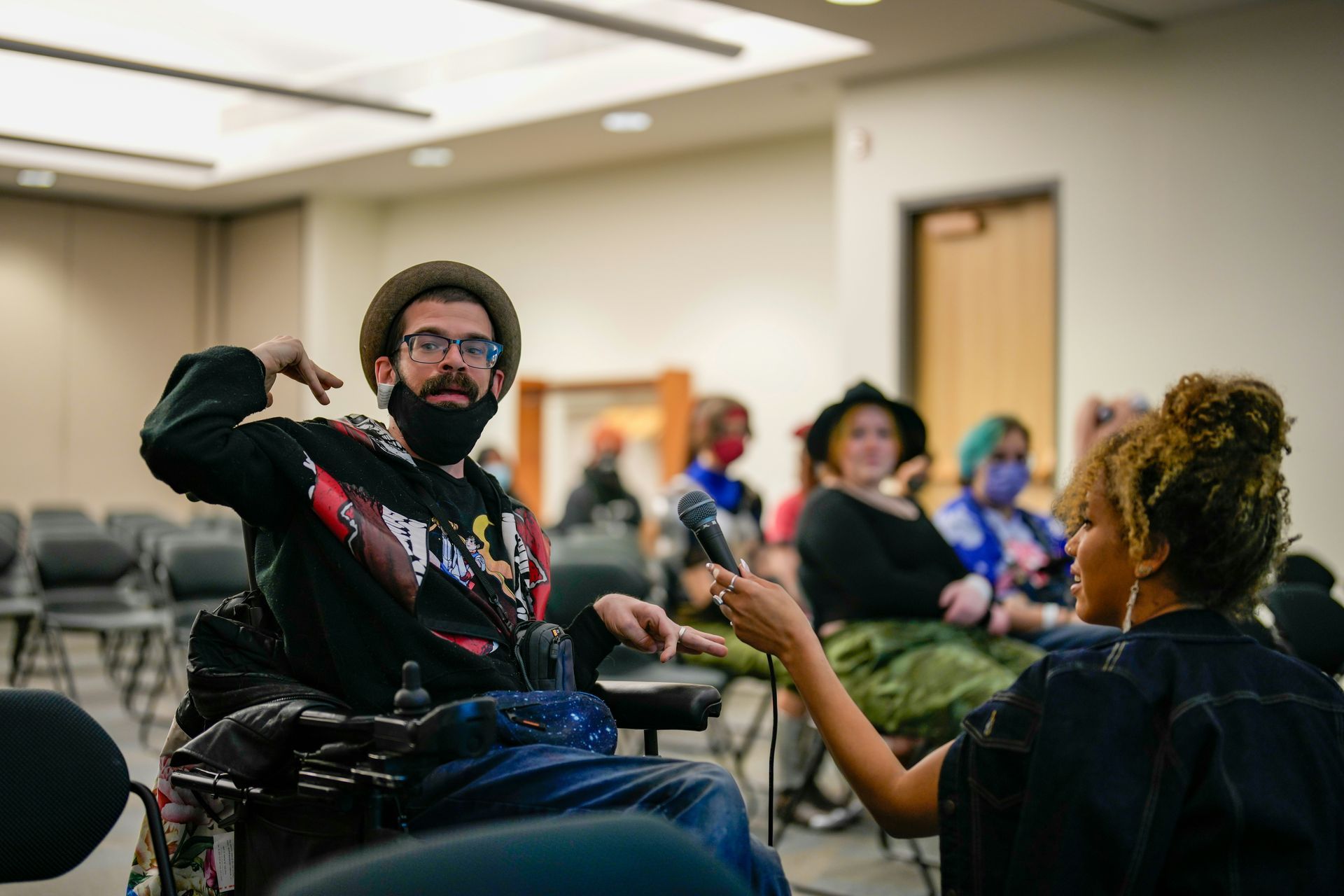 A person in a wheelchair with a hat and mask gestures while speaking into a microphone held by another person indoors.