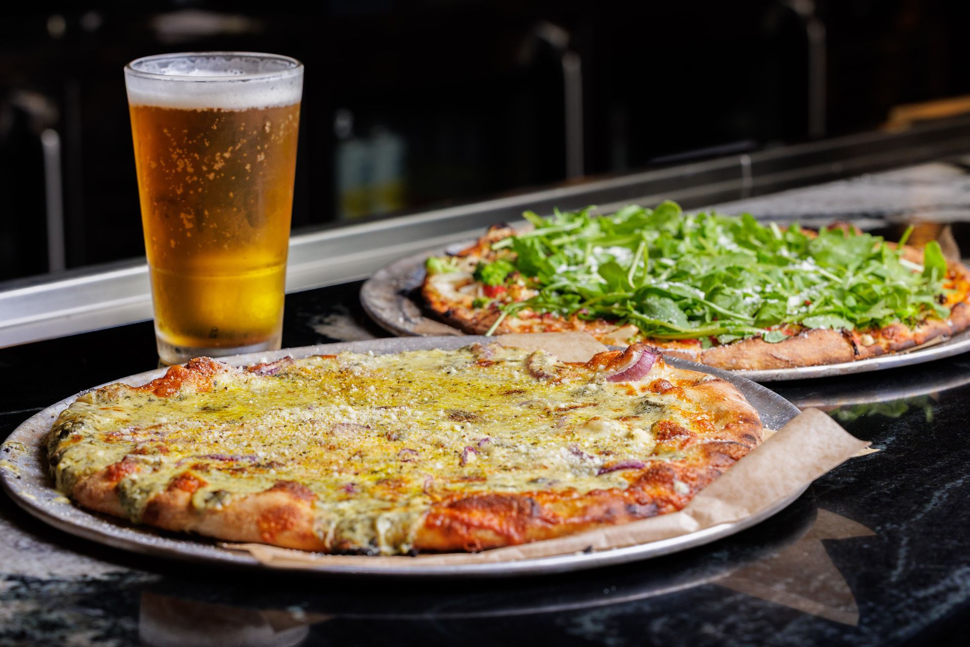 Two pizzas and a tall beer on a table, one cheese pizza and one topped with greens at Brewbury
