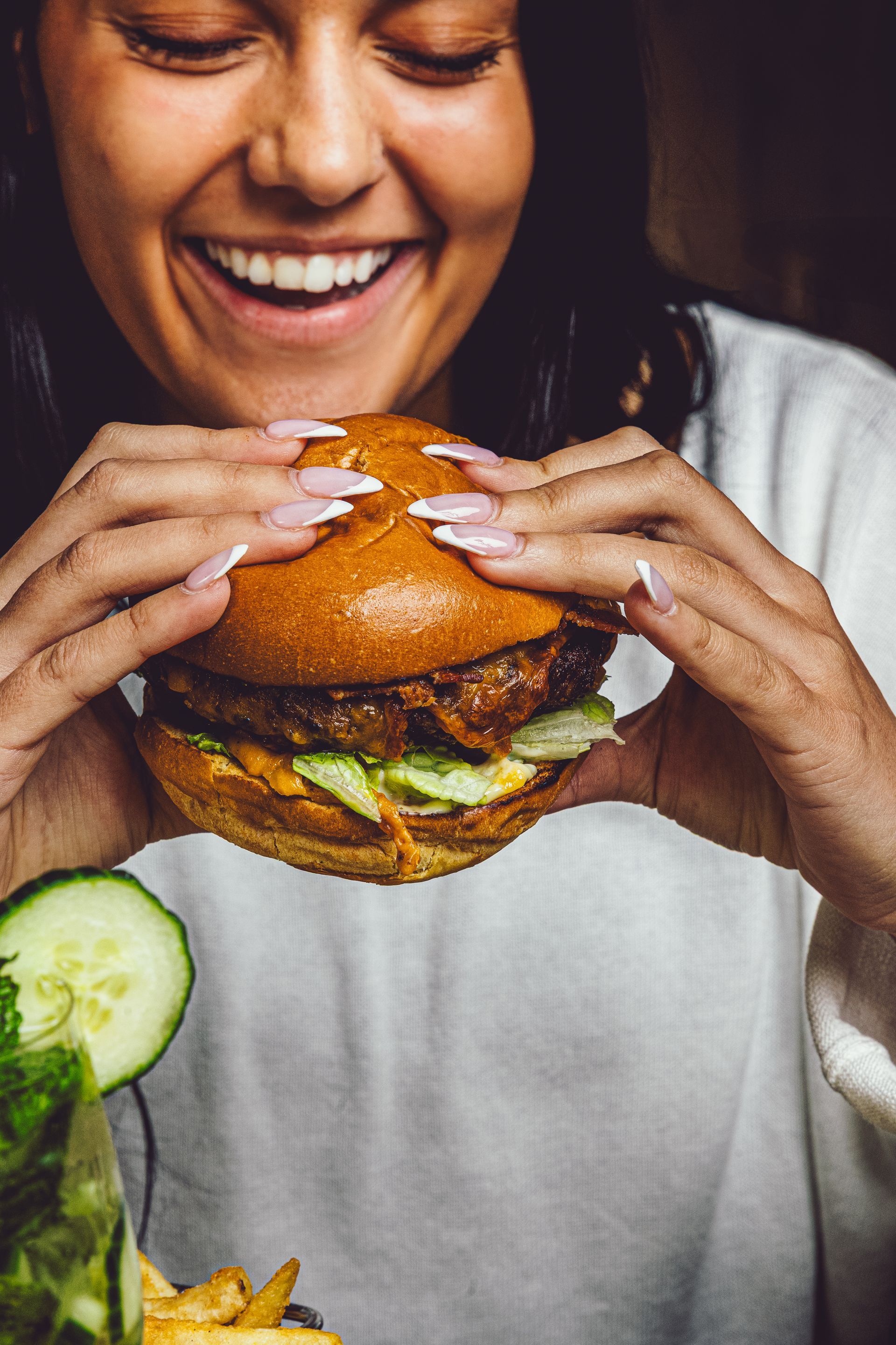 Smiling person holding a large burger with a cucumber slice in the foreground