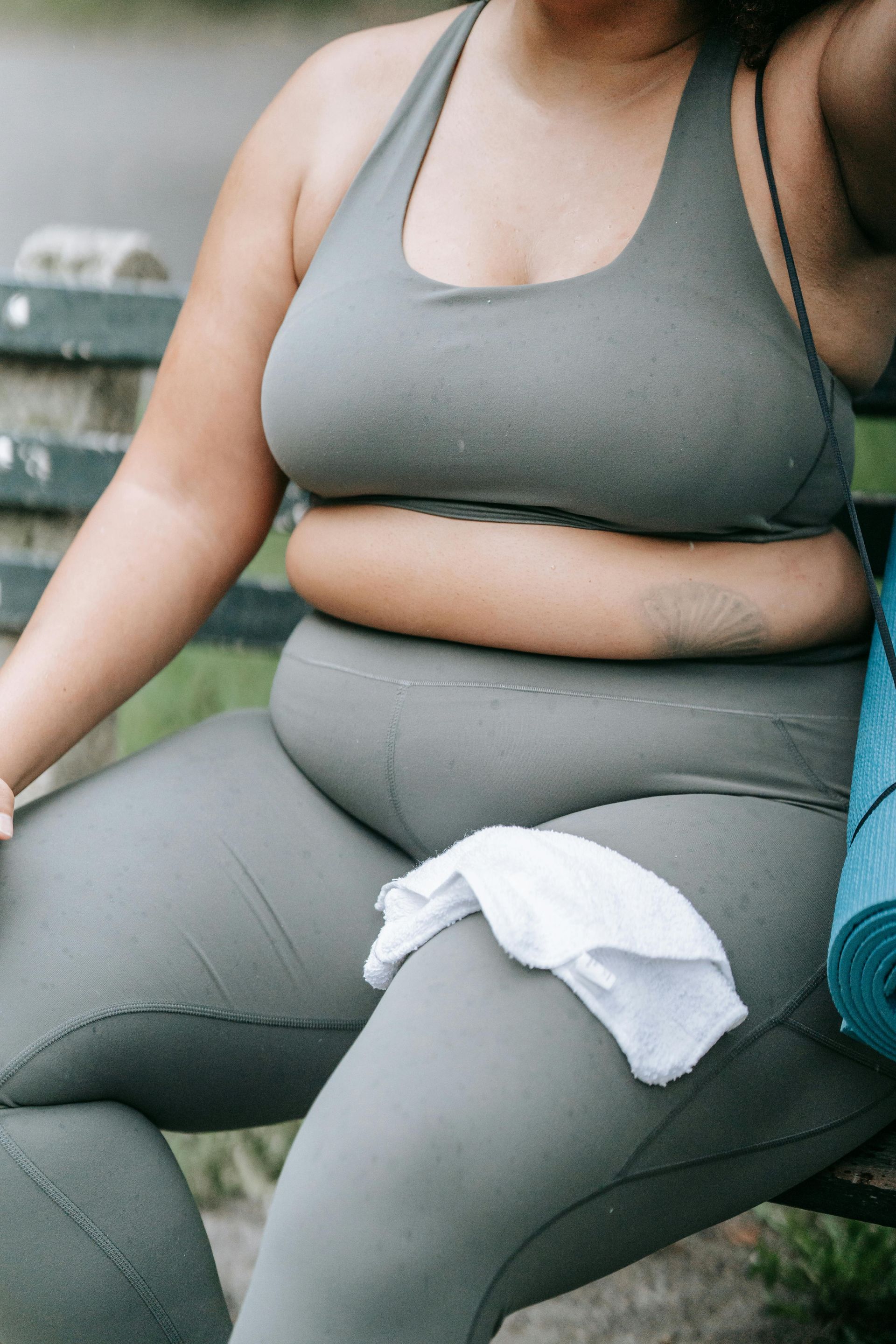 Kvinna i grå träningskläder sitter på en bänk med en handduk och yogamatta.