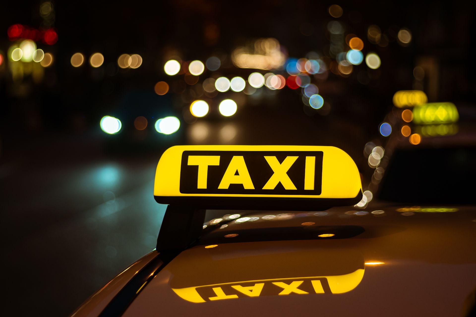 Gul taxiskylt på biltaket på natten med suddiga stadsljus i bakgrunden. Läkarintyg krävs för Taxikörkort