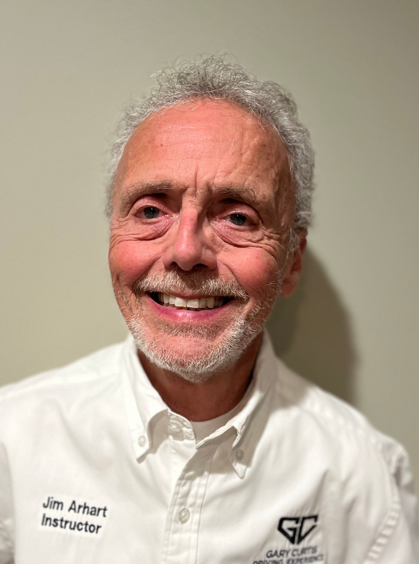 Smiling man, wearing a white shirt with text, appears to be an instructor, against a neutral background.