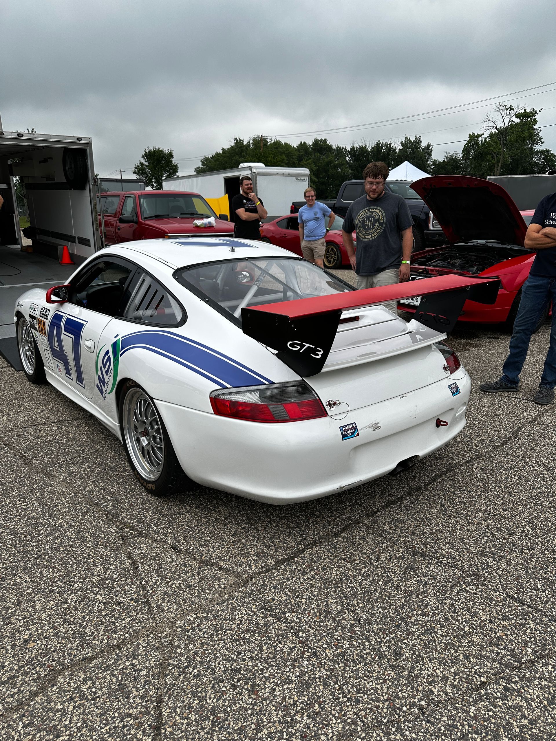 White race car with blue accents and a red wing parked on gravel at a car event, with people and trucks nearby