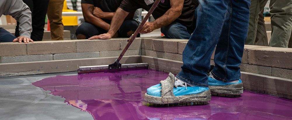 Person squeegeeing purple paint on pavement beside stacked bricks, wearing blue sneakers.