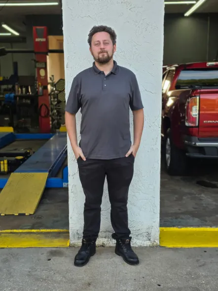 Man standing in a garage doorway beside a red truck, wearing a gray polo and black pants.