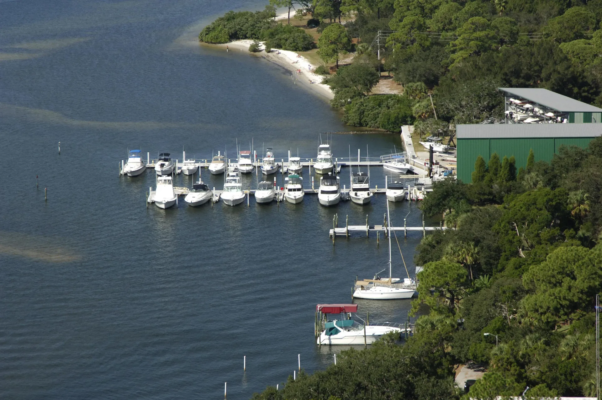 Several white boats docked on a calm blue bay beside a green, tree-lined shore